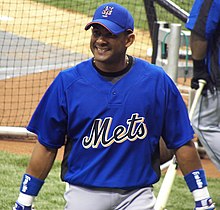A man, wearing a blue baseball cap with an interlocking "NY" at the center, a blue baseball uniform with the words METS across and a blue wristband around both wrists, smiles as he takes batting practice