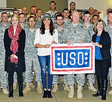 Dianna Agron and Meghan Markle, dressed casually and surrounded by military service personnel, hold a USO banner.