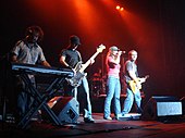 Four people on a stage. The first one with a plaid shirt and blue jeans, playing a keyboard. The second one wearing a black cap, a striped shirt and blue jeans, playing a bass guitar. The third one is a female wearing a gray hat, a pink shirt and blue jeans, holding a microphone. The fourth one, is wearing a gray shirt and blue jeans and is playing an electric guitar.