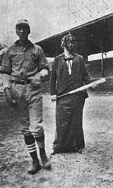 A man and a woman standing on a baseball diamond. The man is wearing a player's uniform; the woman is wearing a loose, tailored suit and an umpire's mask, and holds a bat.