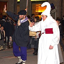A night scene. A woman in white with an elaborate headdress walks hand-in-hand with a man in a dark Basque farmer outfit.