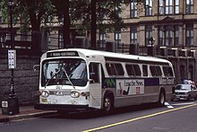 Old film photo of an older-model GM high-floor Metro Transit bus running southbound on Barrington Street next to the tall stone retaining wall of the Grand Parade. The bus is painted with a wide blue stripe down its length, the words Metro Transit in white at the rear of the bus. The photographer seems to be standing in front of the TD Building. Halifax City Hall is visible in the background.