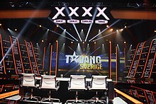 Photograph of the setup showing 4 chairs for the judges and four X-shapes above them with their names, and in the background there seems to be a stage