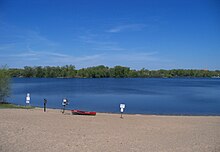 A beach along Cedar Lake with a red and white water safety boat on its sand. Lush green trees line the opposite shore across the deep blue water. A danger sign sticks out of the sand warning people to stay off the lake when it is frozen.