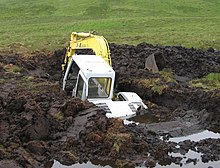 Photograph of a backhoe that is over half submerged in a large hole that it dug in a peat bog before falling in.