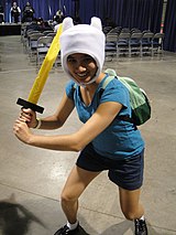 A young woman dressed up as Finn the Human, wearing his white bear hat, blue shirt and short, and green backpack while wielding a sword