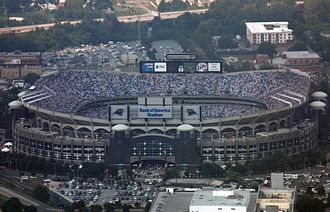 Aerial shot of an open-air stadium during a football game. The outside facing is defined by a series of arches, and scoreboards are visible at the top of the facility.