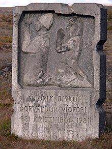 A rectangular stone marker with Romanesque-style bas relief depicting two men knelt in prayer. Text below the relief reads: Fridrik Biskup – Þorvaldur víðförli – 981 Kristniboð 1981