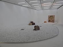 An exhibition gallery, with white pebbles on the floor, five images on slabs and a small block of sandstone to the back.