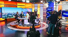 A look behind the scenes at a news set. Several people are seated at the desk. Two large cameras are visible, with a director standing between them.