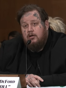 Jelly Roll, dressed in black, sits and speaks earnestly into the microphone in formal proceedings