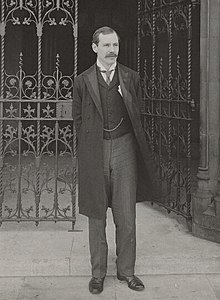 Standing portrait photograph of Arkwright, in jacket and waistcoat, outside some wrought iron gates
