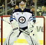 An ice hockey goaltender stands in front of the net. He wears white leg pads and glove and blocker. He is wearing a white jersey with blue shoulders with a jet over a maple leaf inside of a circle for the logo.
