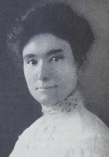 A white woman with dark hair, eyes, and brows. She is wearing a high-collared white lace blouse, and her hair is in an updo.