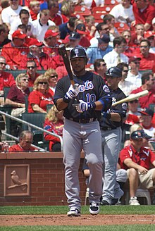A man wearing a black baseball jersey with "Mets" written across the chest, gray pants, and a black batting helmet holds his baseball bat as he approaches home plate to begin his at-bat