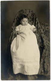 Baby in a long white dress at on what appears to be a chair draped in fabric.