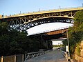 A pedestrian walkway below a car and train bridge, and slightly elevated above a river. It's wide and has a railing on each side