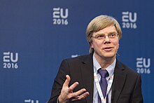 A man with light hair and glasses speaks at a conference. He is dressed formally and is gesturing with his hand. In the background is a blue backdrop with logos reading "EU 2016".