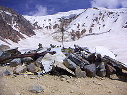 The snowy peak is pictured in the distance with a mound of rocks, debris, and a cross in the foreground commemorating the victims