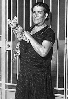 B&W portrait photograph of a standing, middle-aged woman, wearing a dress, holding a cat.
