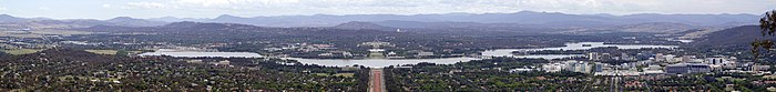 Panoramic view from a mountain overlooking the lake from north to south. View is Axially along Anzac Parade and Parliament House is directly opposite. A group of office blocks and small skyscrapers can be seen at right, the city centre.