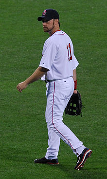 A bearded man in a white baseball uniform and dark baseball cap looks over his left shoulder.