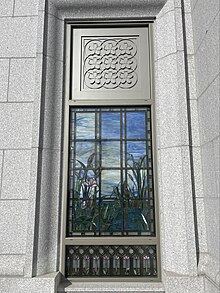 Stained glass depicting various water elements and bullrushes, against a blue sky background. A metal frame outlines the glass and above the stained glass depicts a pattern of nine cherry blossoms. Masonry from the temple walls hold the window in place.
