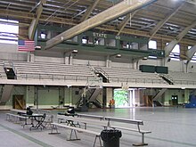 The gym's interior, with a small pressbox and roughly ten rows of bleachers elevated above a grey playing surface adorned with metal benches and a basketball hoop