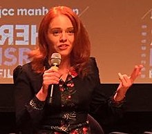Shoulders-up image of Maya Zinshtein wearing a blazer over a floral print dress. She has red hair and is holding a microphone.