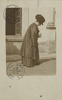 The obverse of a postmarked postcard shows the photographer wearing a dark cape and dress, and hat with plume, leaning over a camera on a street (possibly in Paris).