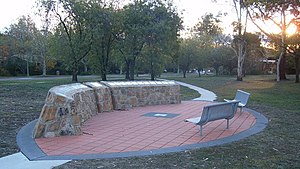 An ellipse of orange pavers surrounded by a grey border, with a man-made rock structure on one side of the ellipse displaying plaques. On the other side are two bench seats. Grass surrounds the ellipse and trees can be seen in the background.