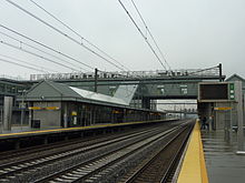 A train station with overhead lines and four tracks between two sheltered platforms, both wet, under cloudy skies. Letters across an overhead walkway spell out "Newark Liberty International Airport"