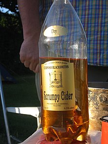 A plastic bottle of dark gold liquid sitting on a table. A gold label reads "Scrumpy Cider".