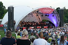 The orchestra on an open-air stage under a vaulted white tent roof; in front of it a large audience.
