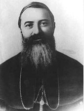A bearded man wearing a pectoral cross around his neck faces forward.