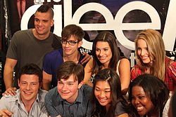 A group of people huddled together, with the backdrop displaying the word "Glee" in white small fonts.