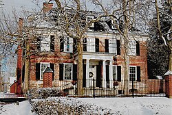 Red brick building with snow on the dark roof. In front of the building, there are trees and snow on the ground.