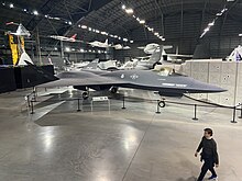 The YF-23 painted in charcoal gray, the "Gray Ghost" is seen front and center, with a man walking in front of it at the bottom right of the image. Multiple other aircraft held at the museum can be seen behind it