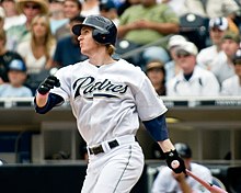 A man in a white baseball uniform with "PADRES" on the chest stands with a baseball bat in his left hand as if having just taken a swing.