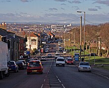 Ina suburban area, a busy main road wit vehicles travelling in both directions recedes into the distance. On the left is a row of red-bricked two-storey houses. On the right, an open parkland fringed with mature trees is visible. In the distance is a sunlit landscape of urban and industrial buildings leading to distant, low hills.