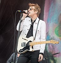 Britt Daniel, wearing a white denim jacket, sings into a microphone