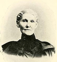 An older white woman wearing black, with white hair parted center and drawn back.