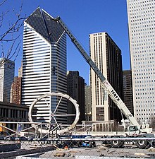 A crane lifts a large, oval-shaped ring in front of several large buildings