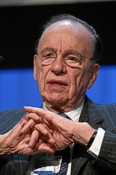 A spectacled man in his seventies faces to the left of the viewer. His hands are in front of him, the fingers interlinked.
