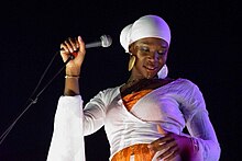 An image of India.Arie performing in a white dress and head wrap. She is looking away from the microphone.