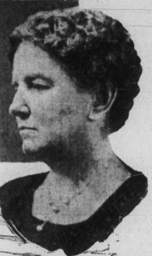 A middle-aged white woman with short curly hair, wearing a dark top with a round neckline