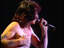 Scott, aged about 33, is shown in right profile, he holds his microphone in his left hand, while his right hand, with tattoos visible, is clenched near his bare chest.