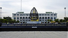A granite sign with a long name in Thai script, and a building in the background