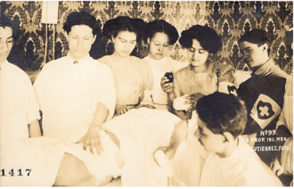 A sepia photograph depicting six doctors and nurses standing in front of a tapestry and observing a seated doctor on the right caring for a patient lying on an operating table.