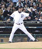 Man in a white baseball uniform stretches to throw a baseball with his left hand